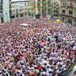 Se puede ver la plaza del ayuntamiento de Pamplona en pleno 6 de julio llena de personas. Dos tercios de la imagen los ocupan pequeñas cabezas tintadas de blanco y rojo. Imagen multitudinaria